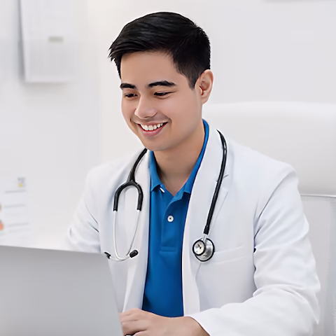 Dr. Yaro Caracas wearing a white coat and stethoscope smiling while using a laptop in a bright medical office